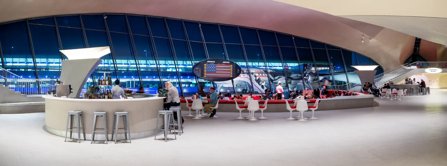 Sweeping white interior of the TWA Flight Center at night, with red Sunken Lounge seating and Connie outside the glass wall.